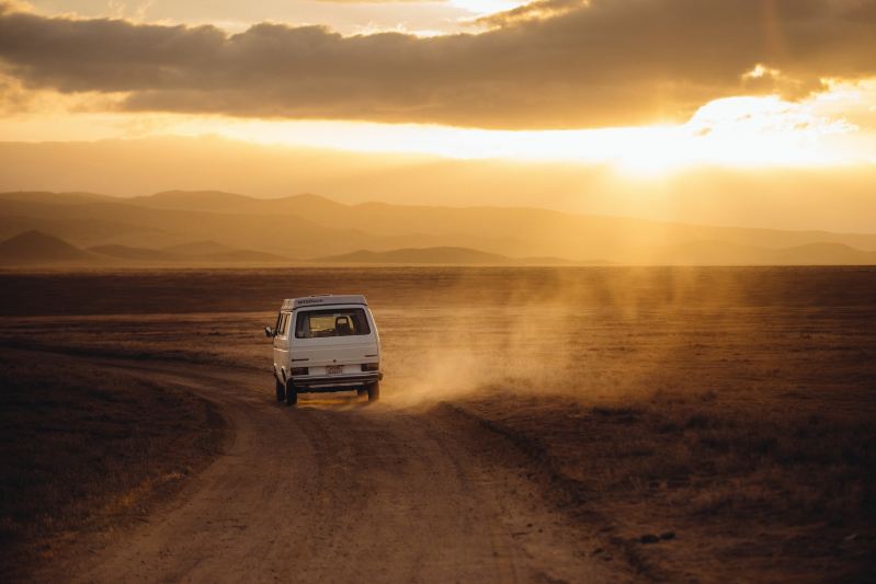 White campervan driving a dusty road in a hot desert landscape.