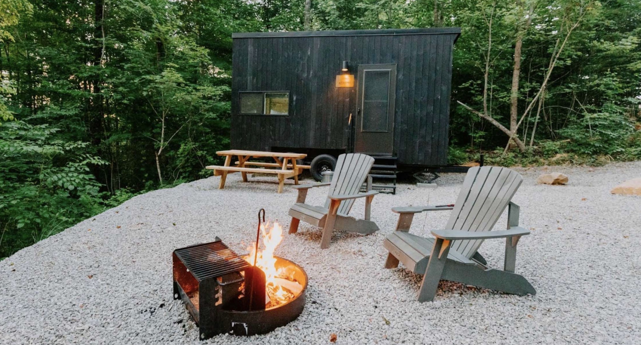 A small, black wood-paneled tiny house in the woods with a firepit and chairs in the foreground