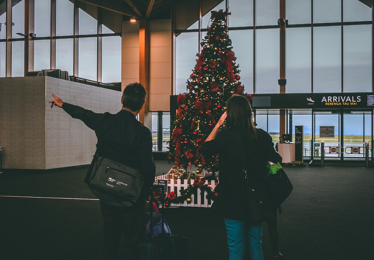 Two travelers with their backs to the camera, gesturing to a large Christmas tree inside of an airport