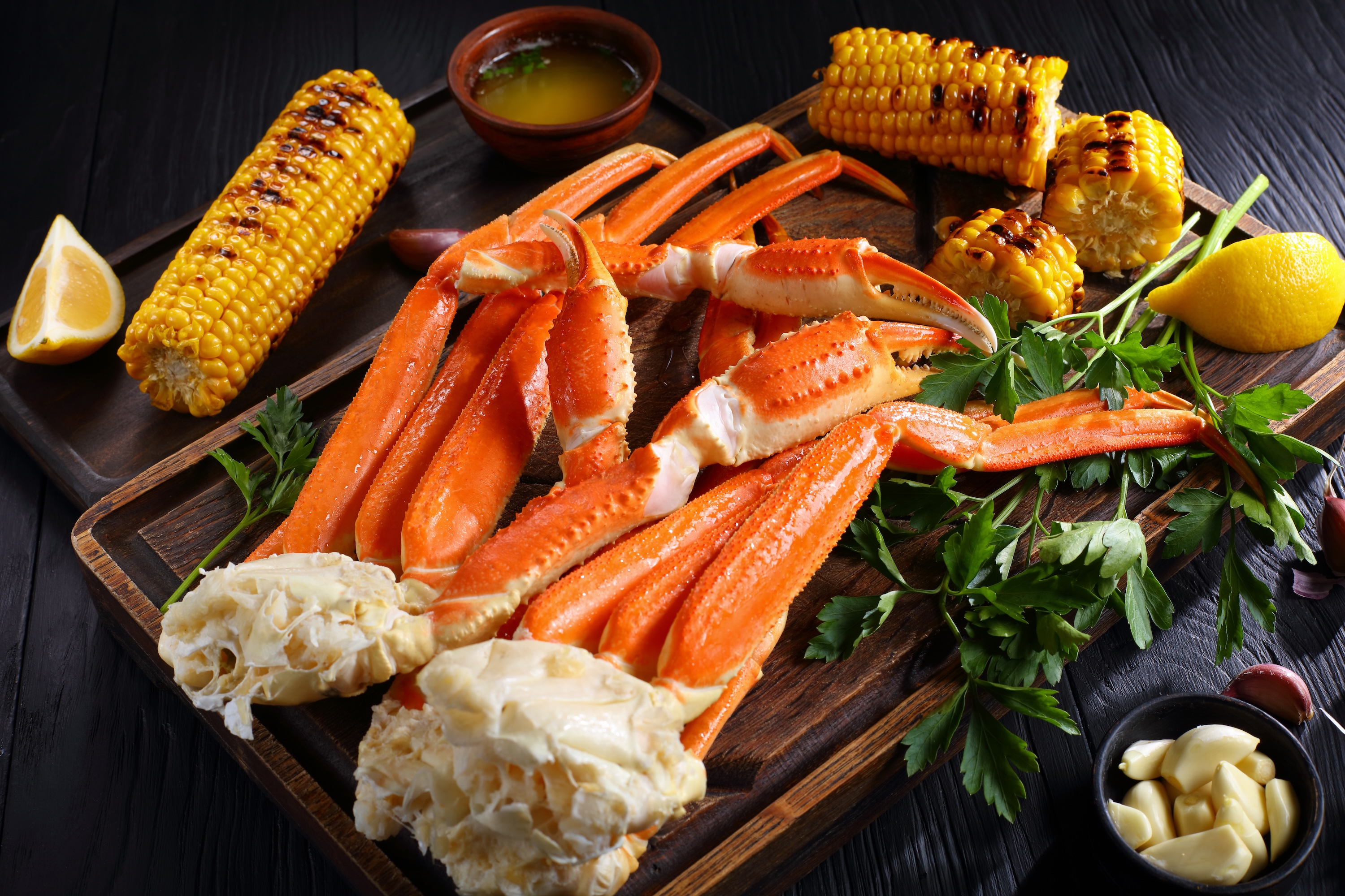 Snow crab legs served with melted butter, garlic cloves, lemon slices, grilled corn in cobs and fresh parsley on wooden cutting boards, horizontal view from above, close-up