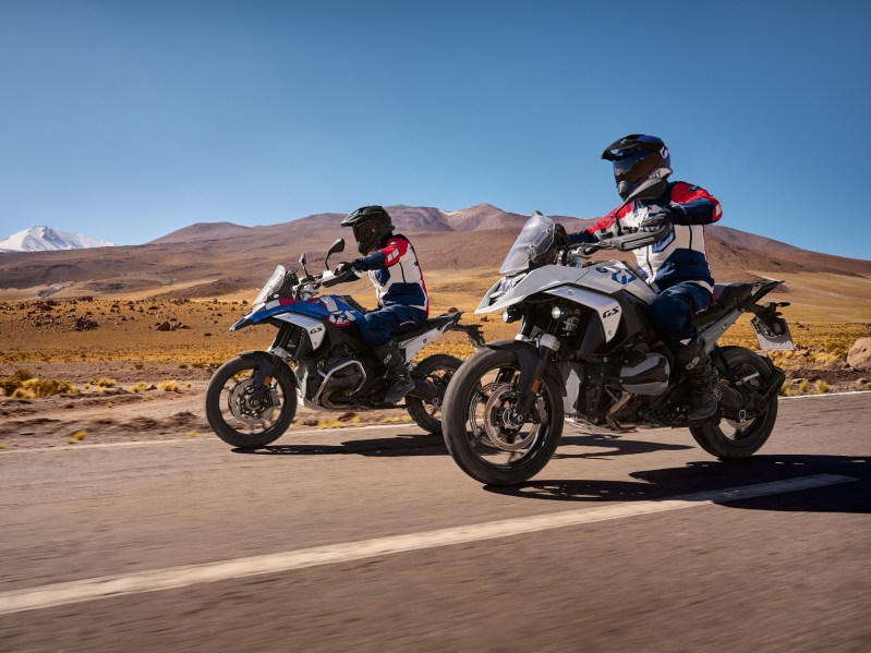 Two 2024 BMW R 1300 GS adventure touring motorcycles traveling on a highway with desert and distant mountains in the background.