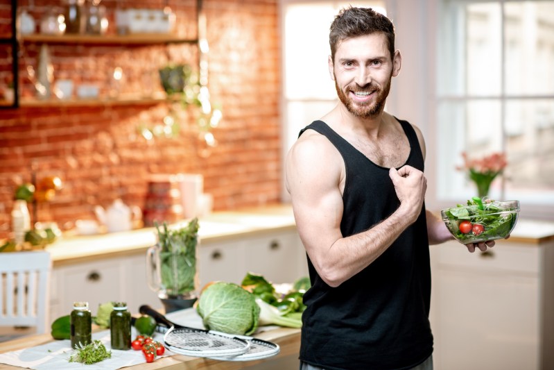 Portrait of a handsome sports man showing muscles, eating healthy vegetarian salad on the kitchen at home