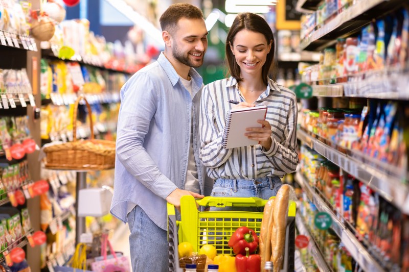 Portrait Of Two Happy Customers Holding Grocery Shopping Checklist, Writing In Notebook, Buying Products, Standing With Full Shop Cart In Supermarket. Young Millennial Family Choosing Healthy Food