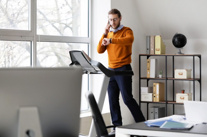 Young man talking by mobile phone while running on treadmill in office