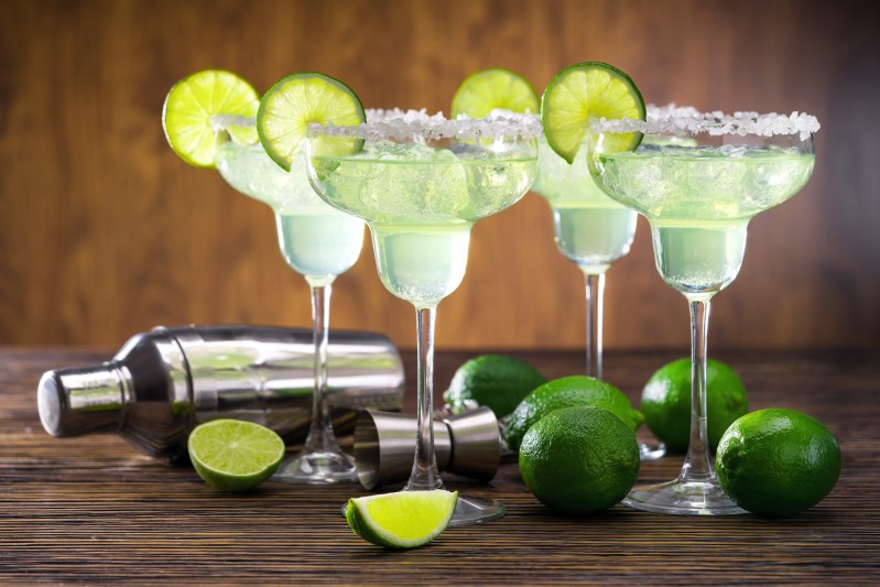Four glasses of freshly made Margarita cocktail decorated with limes on wooden table with shaker and limes.