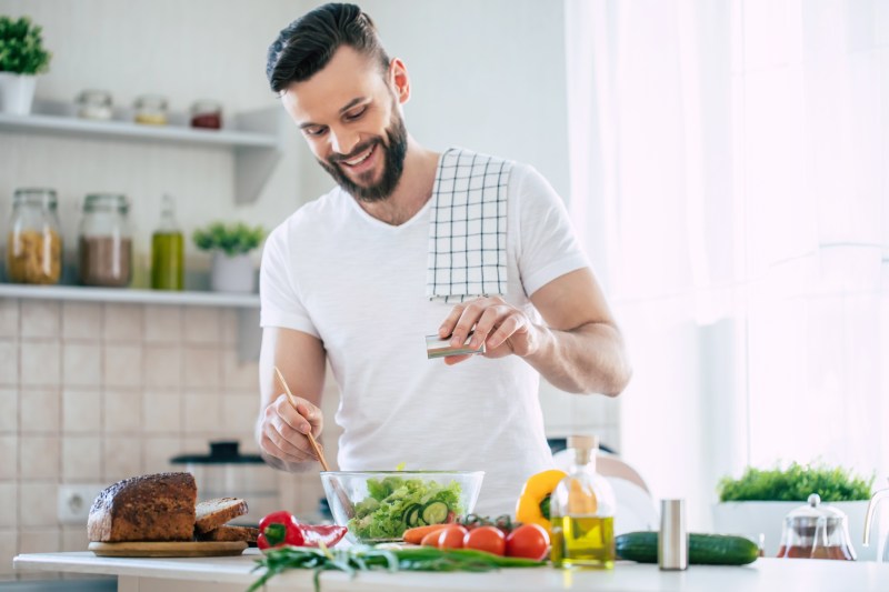 Handsome happy bearded man is preparing wonderful fresh vegan salad in the kitchen at home