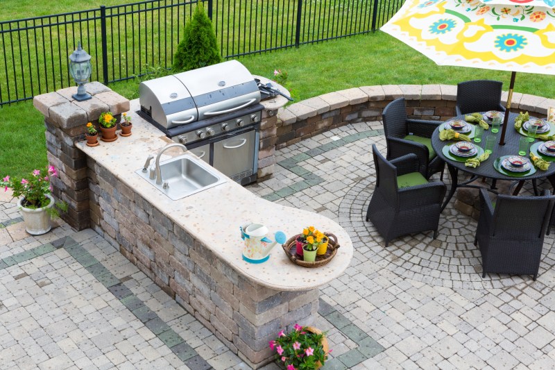 High angle view of a stylish outdoor kitchen, gas barbecue and dining table set for entertaining guests with formal place settings and flowers on a paved patio