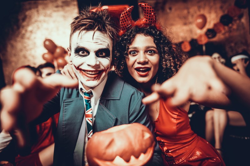 Portrait of Young Couple in Halloween Costumes. Beautiful Woman and Handsome Young Man Wearing Costumes holding Pumpkin at Halloween Party in Nightclub. Happy Friends having Fun Celebrating Halloween