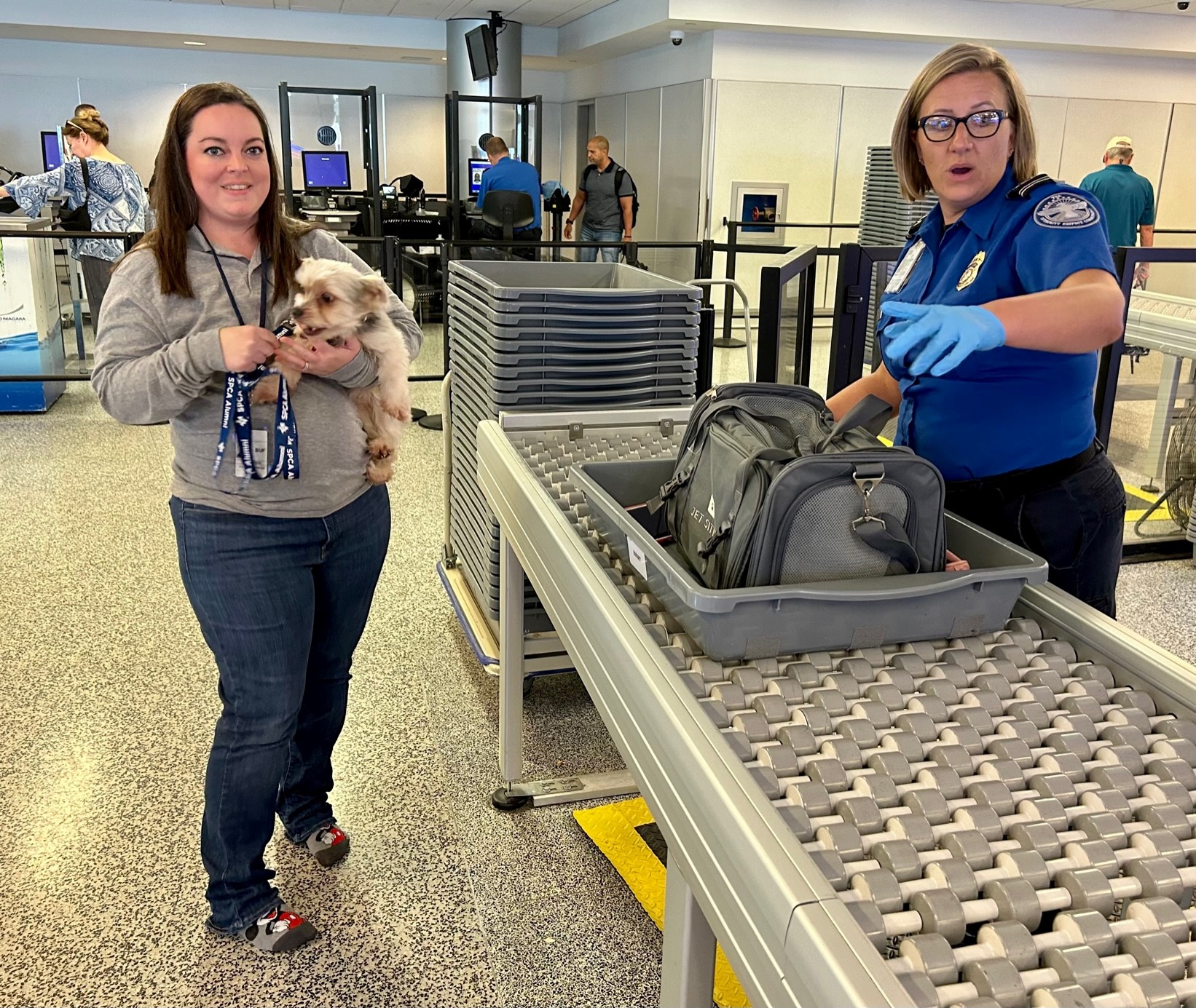 A woman holding her dog while the pet carrier does through the X-ray.