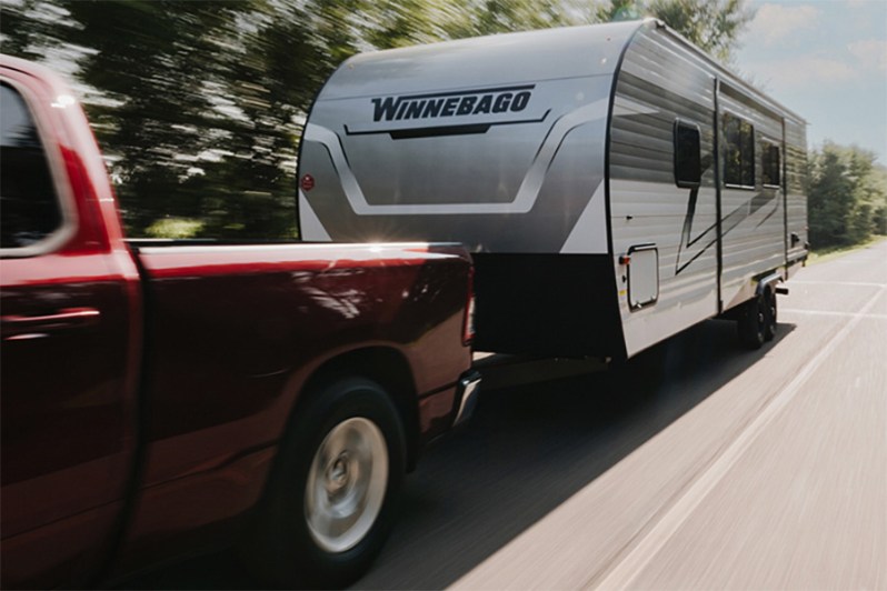 Red pickup truck towing Winnebago Access travel trailer down the road.