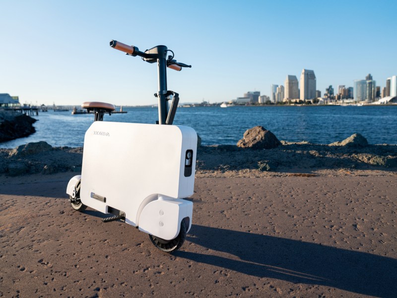 A Honda Motocompacto escooter parked on pavement in the foreground of a large lake with a city skyline in the background.