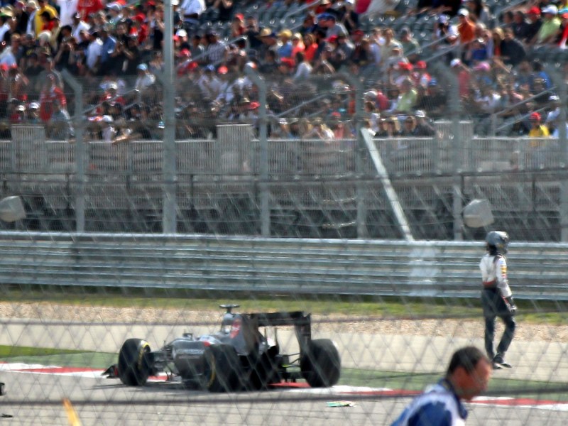 F1 driver walking away from a race car crash in front of a stadium of fans behind fences and safety barriers