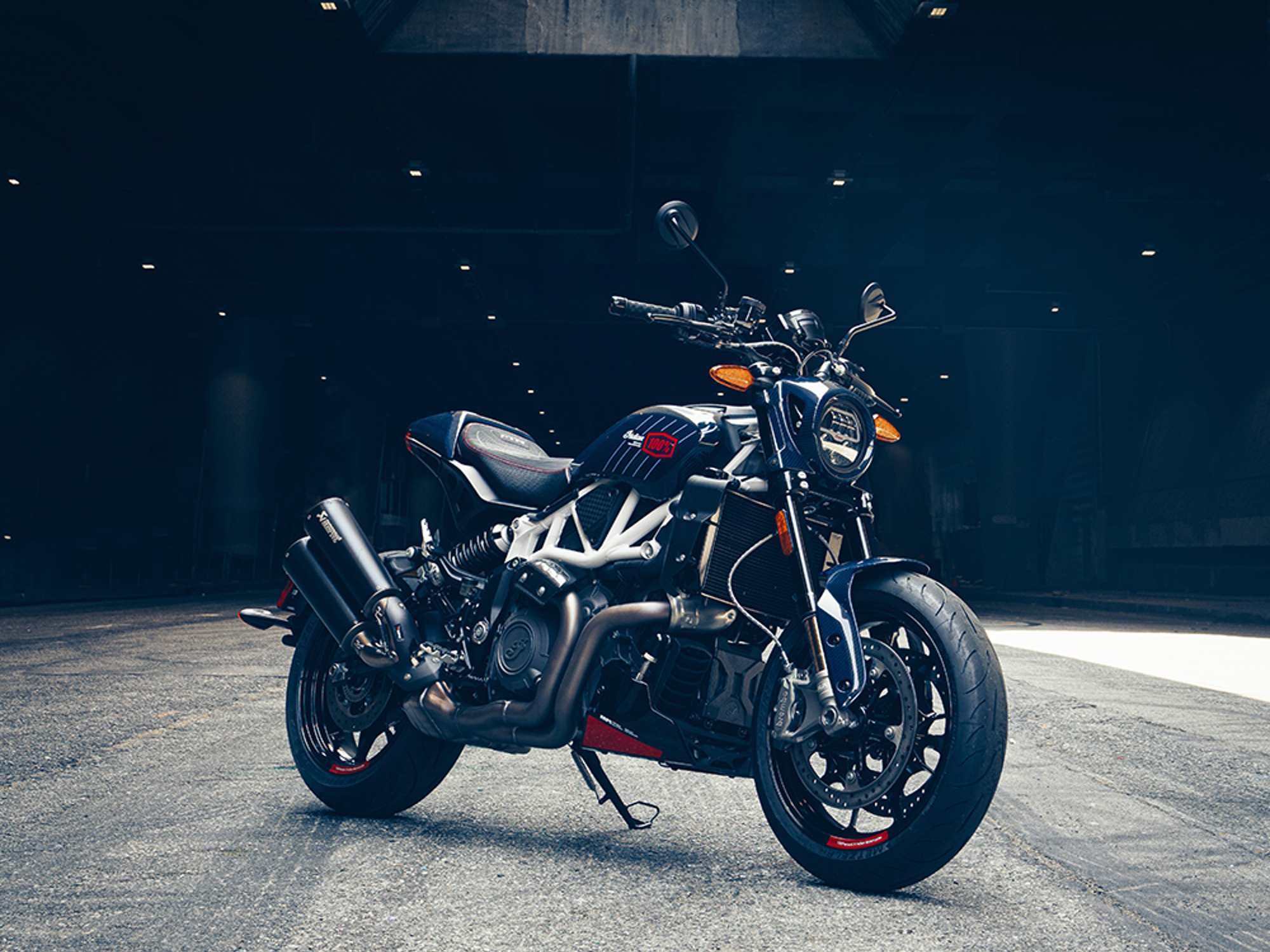 Left front three-quarter view of an Indian Motorcycle 2024 FTR x 100% R limited edition on a light gray floor with a dark background with pin lights