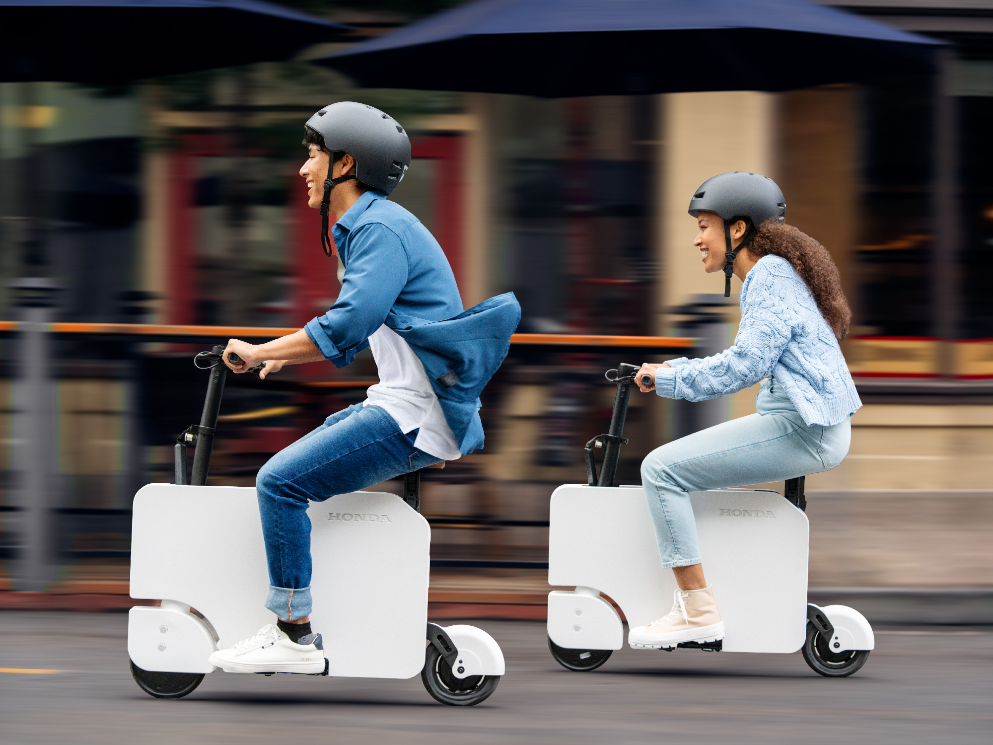 Two people riding Honda Motocompacto escooters on a city street.