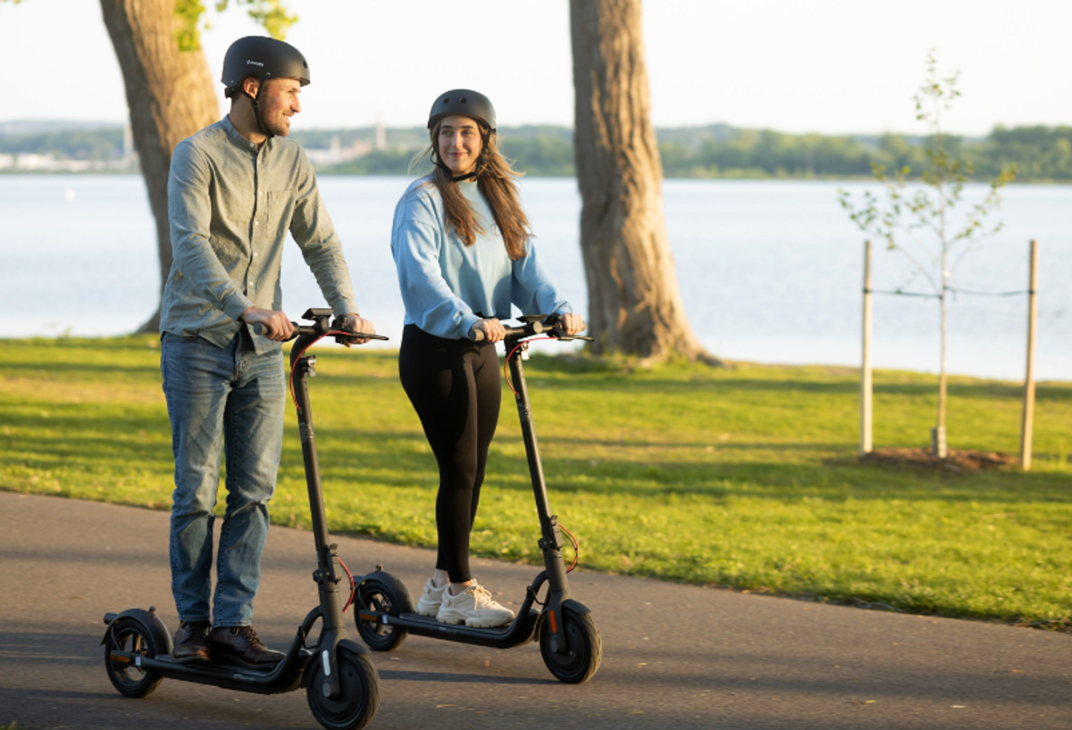 Two people wearing helmets and riding NAVEE V40 Pro electric commuter scooters on a paved path through a grassy area beside a lake.