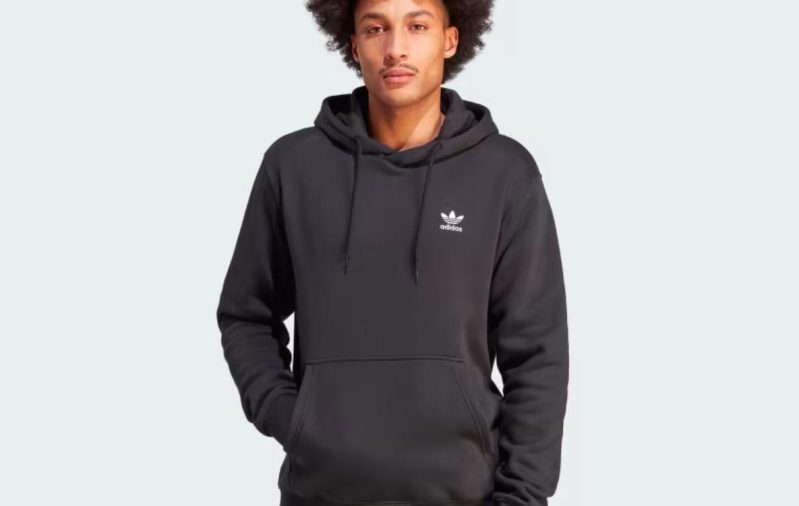 A model wears a black Adidas Trefoil Essentials Hoodie against a gray background.