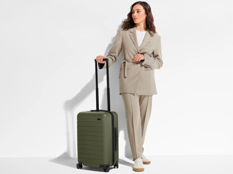 A woman stands next to the Away Travel Carry-On suitcase in olive green.