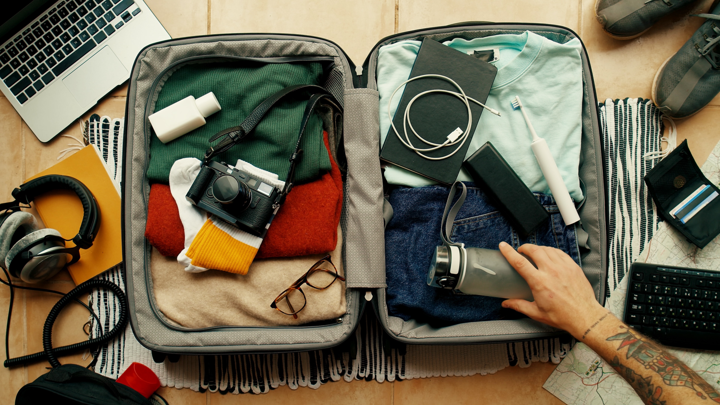 Man with tatted arm packs suitcase for adventure travel trip. Packing bag with camera, laptop, portable battery charger and electric toothbrush.