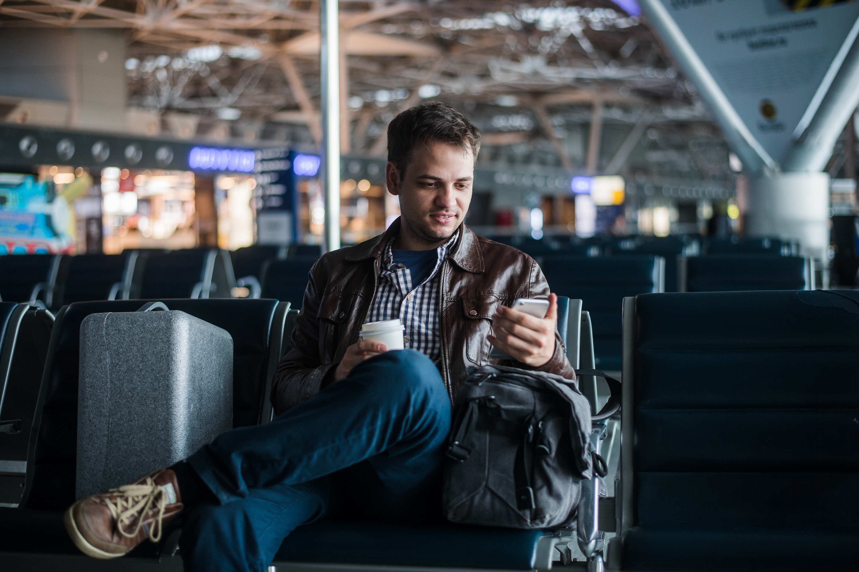 Portrait of handsome smiling man in casual wear holding his luggage and messaging through his mobile phone while sitting in the hall of the airport.