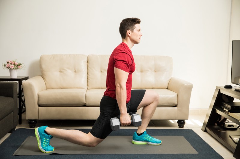 Profile view of a young and fit man doing kneeling lunges with a pair of dumbbells at home