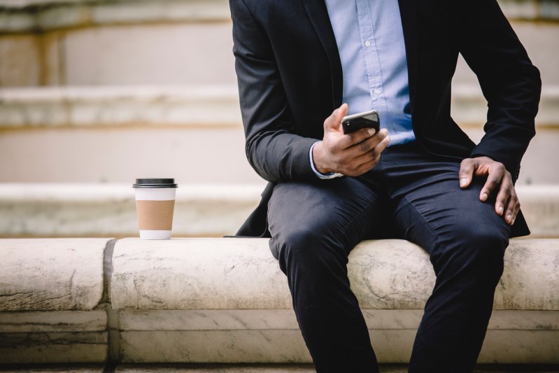 Man sitting next to a cup of coffee on step outside with phone in one hand