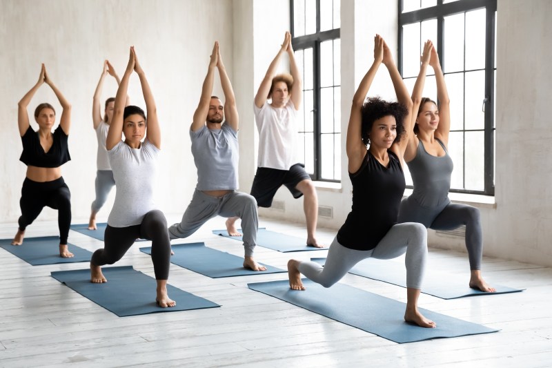 Young diverse young people with African American instructor doing Warrior one exercise, practicing yoga in group class, standing in Virabhadrasana pose, working out in modern yoga studio
