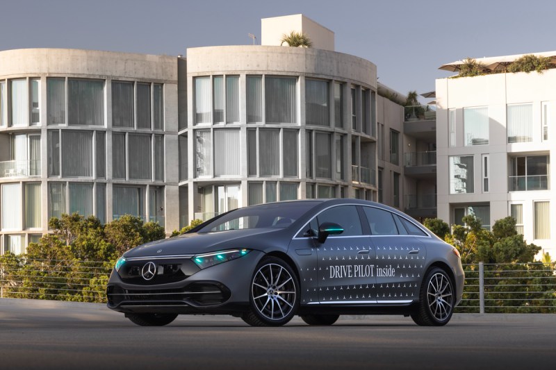 2024 Mercedes EQS equipped with Drive Pilot and turquoise lights left front three-quarter view in front of office buildings.