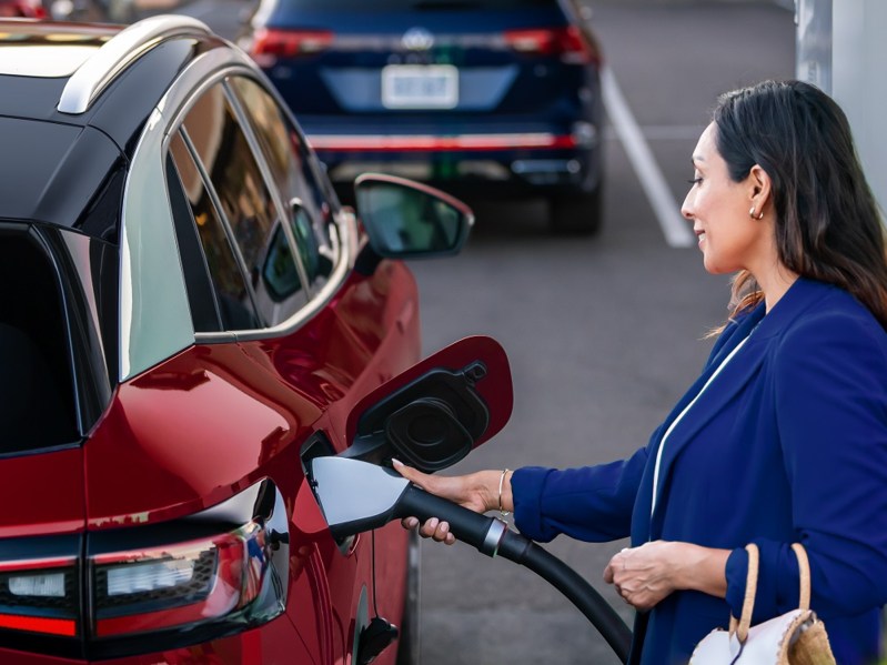 A person standing at the right rear side of a VW ID.4 charging the EV's battery with an NACS charger.