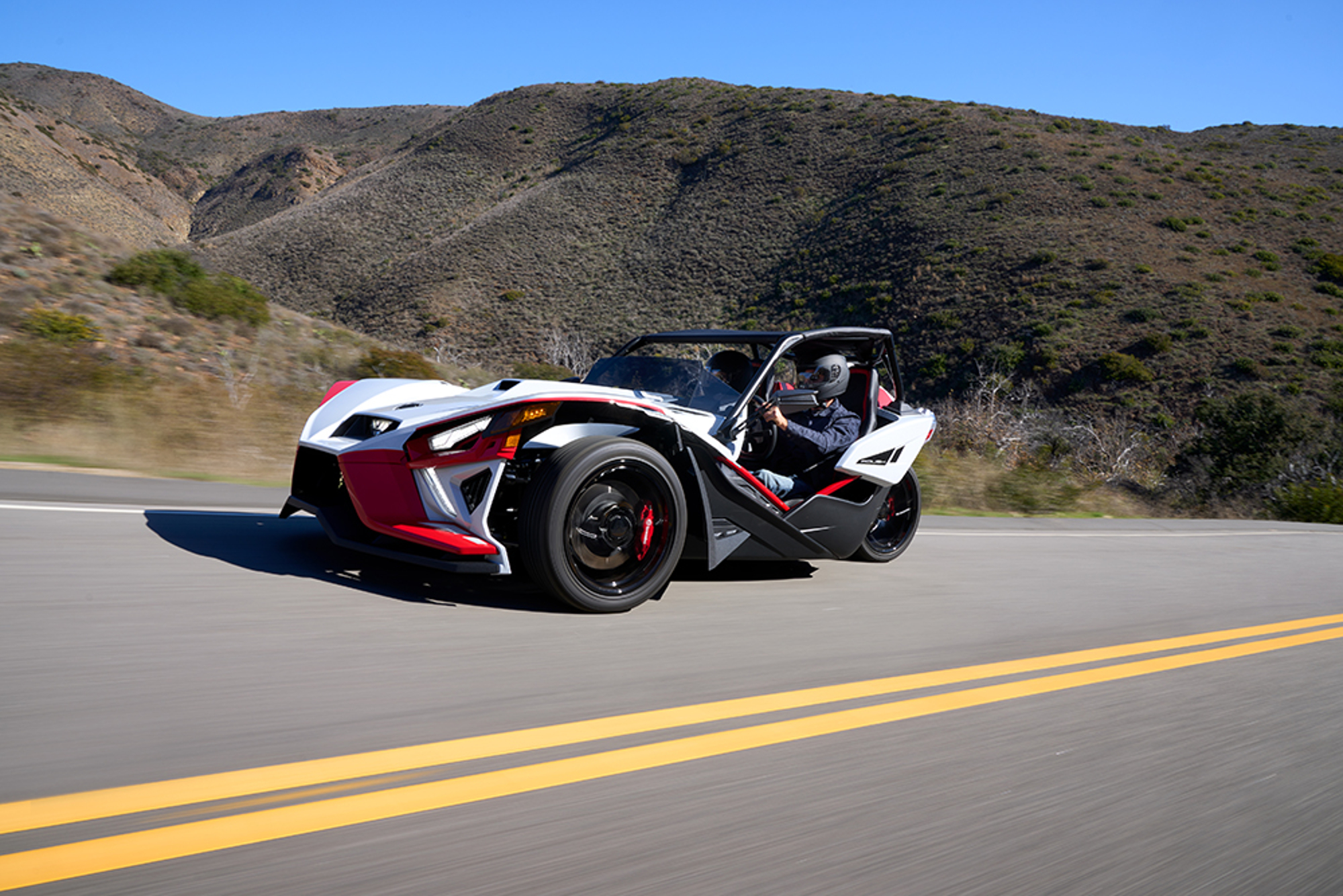 A right front three-quarter view of a 2024 Polaris Slingshot ROUSH Edition with an accessory top traveling on a two-lane road with hills in the background.