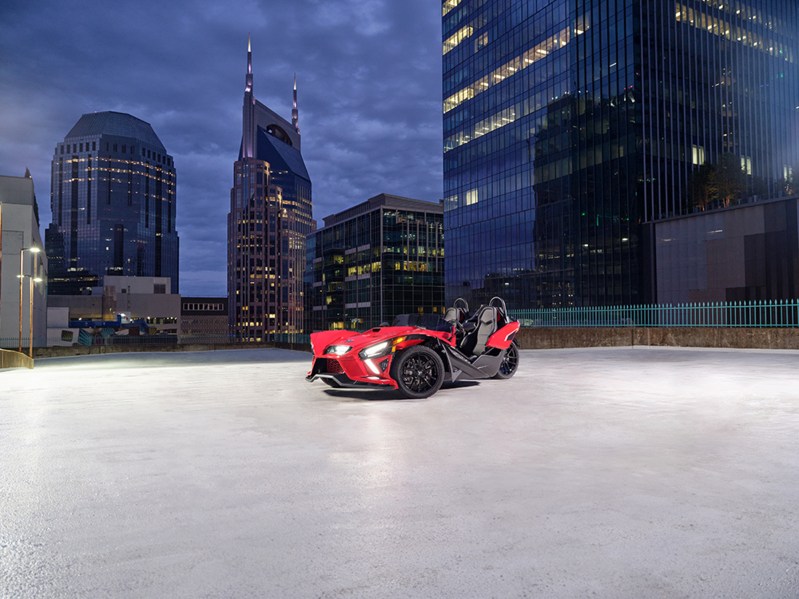 A single red 2024 Polaris Slingshot SLR in an empty parking lot in the middle of a city with large modern office buildings.