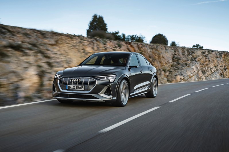Gray Audi e-Tron S Sport left three-quarter view driving on a highway with a rocky ridge in the background.
