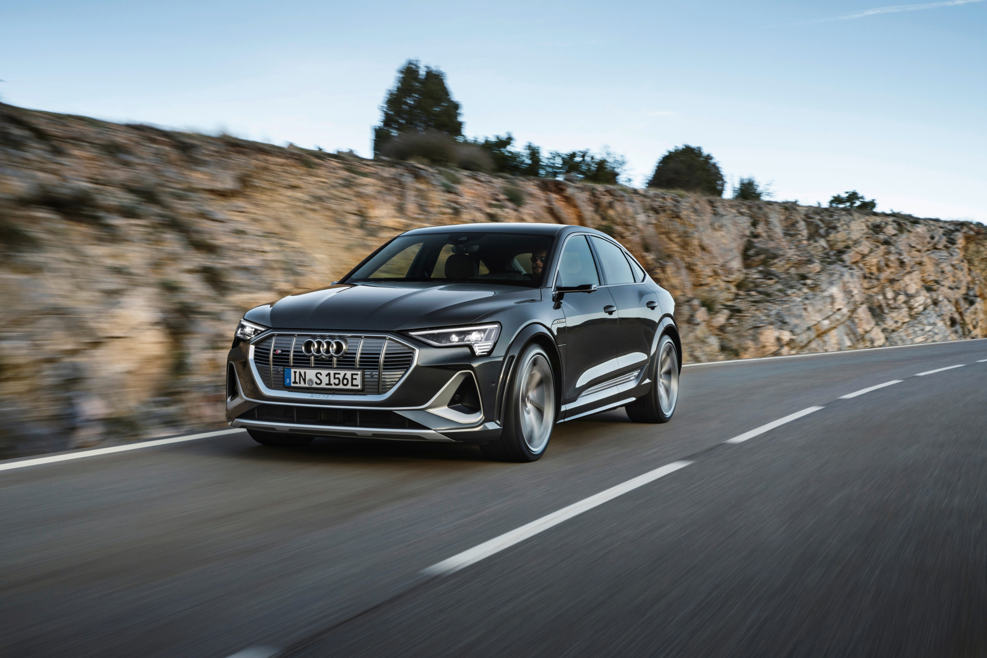 Gray Audi e-Tron S Sport left three-quarter view driving on a highway with a rocky ridge in the background.