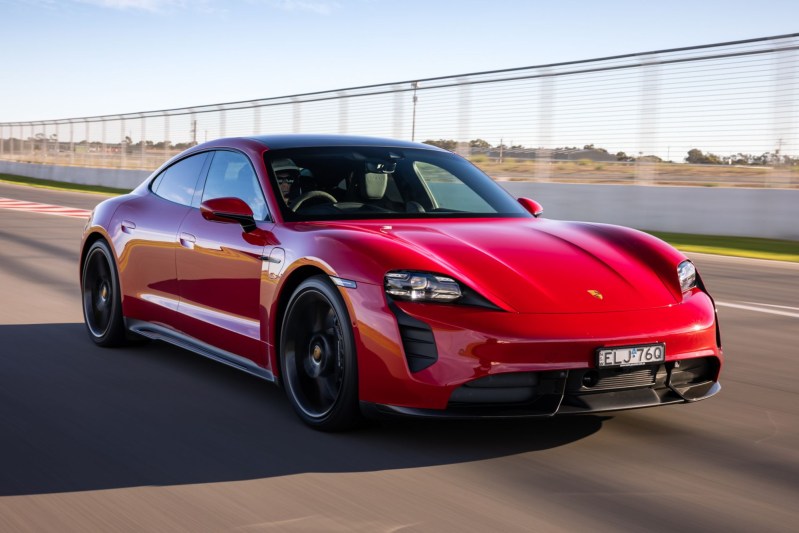 Red Porsche Taycan Turbo S EV right front three-quarter view driving on a highway with a small body of water in the background.