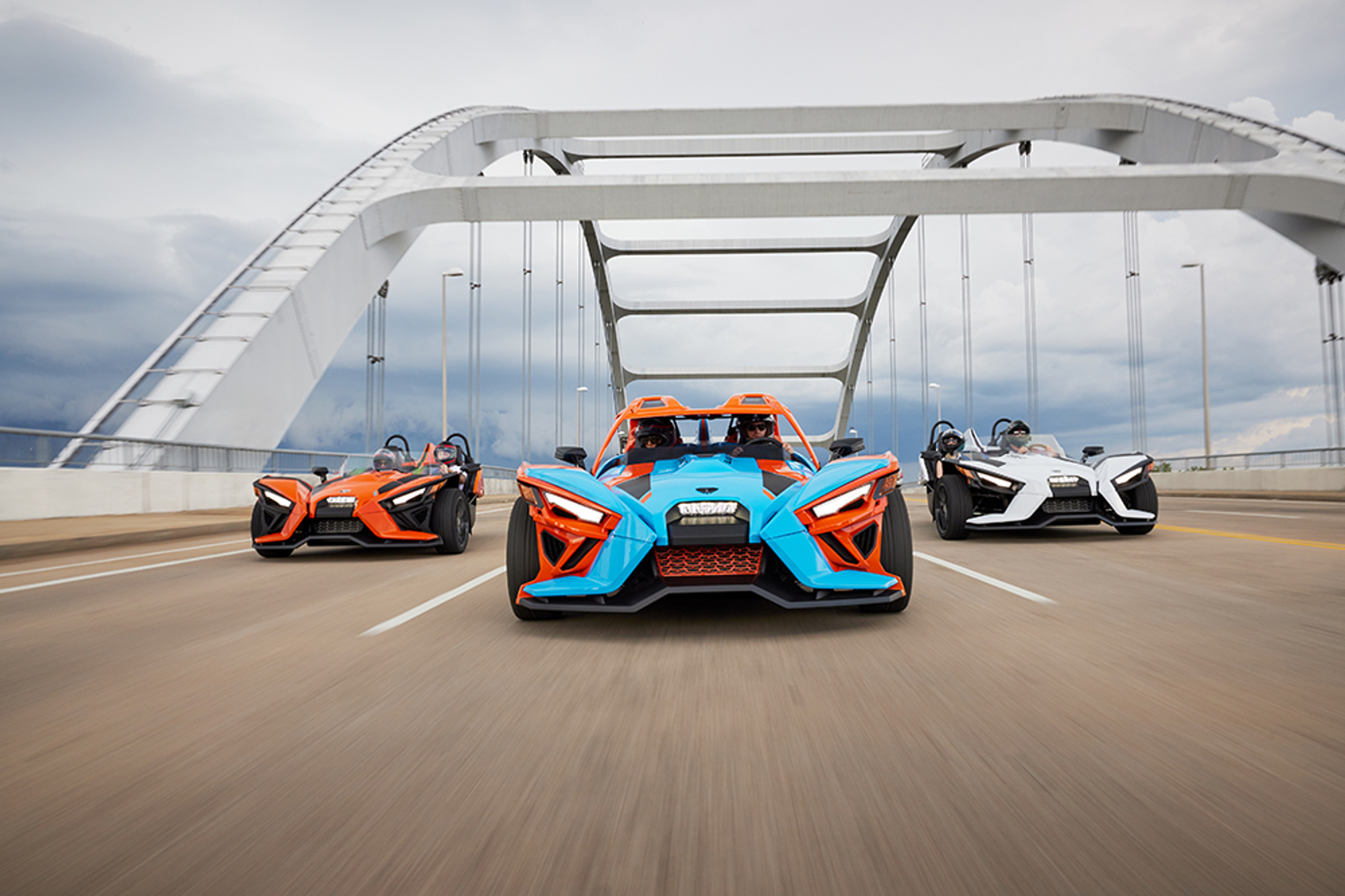 Three 2024 Polaris Slingshot SL, one heavily accessorized coming over a bridge heading drectly toward the viewer.