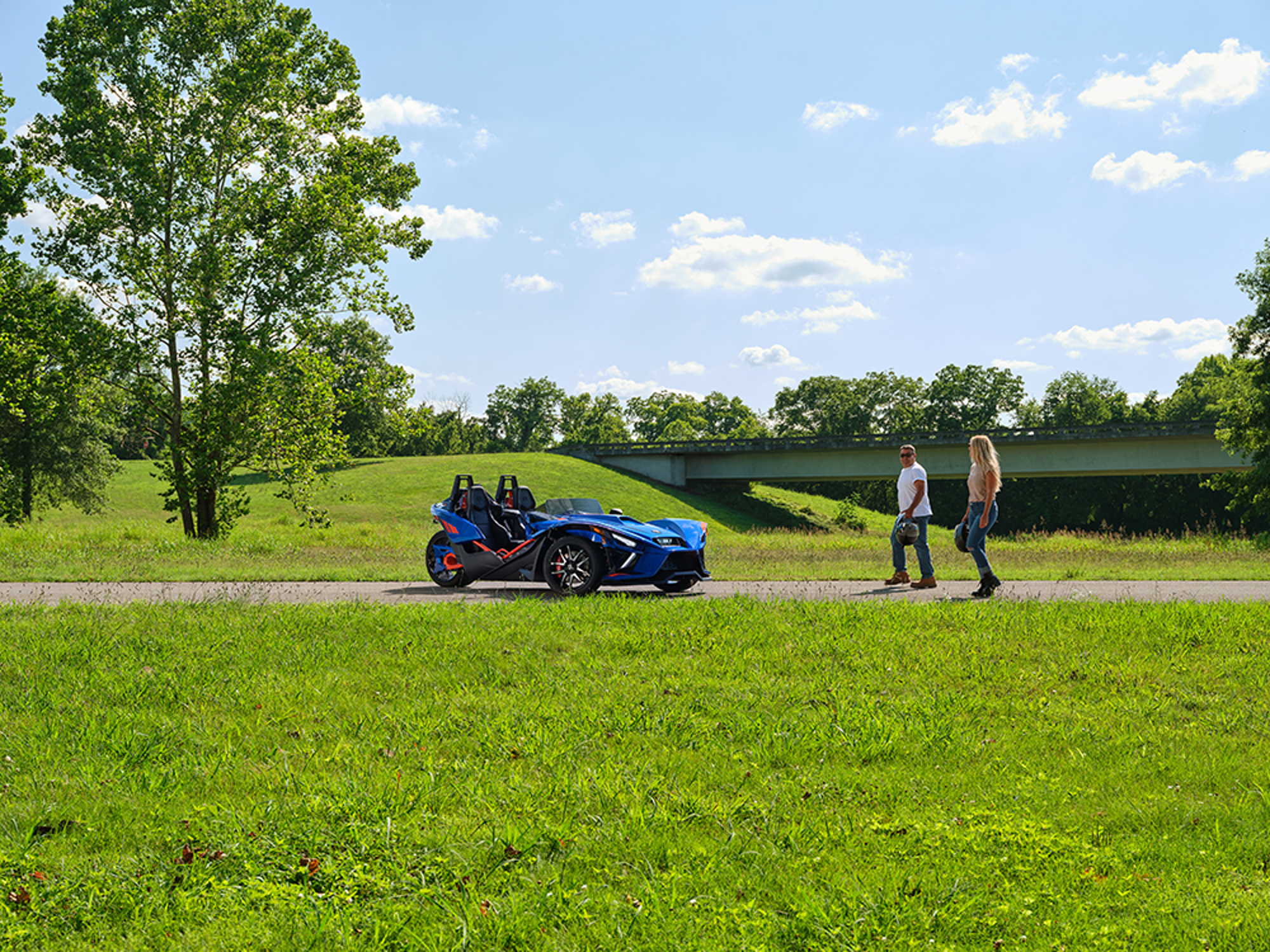 Two people carrying motorcycle helmets walking toward a 2024 Polaris Slingshot R on a paved path through a grassy area with a highway bridge in the background.