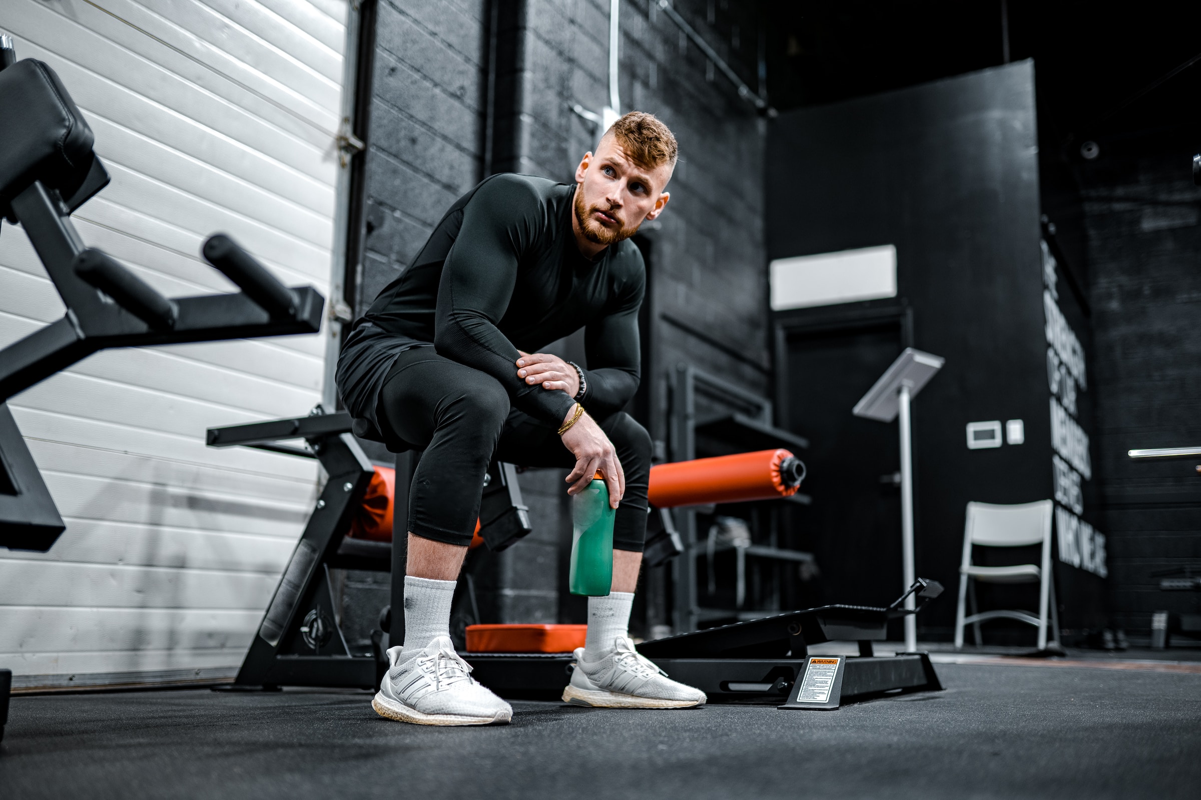 guy sitting on bench at gym holding water flask wearing black with white sneakers