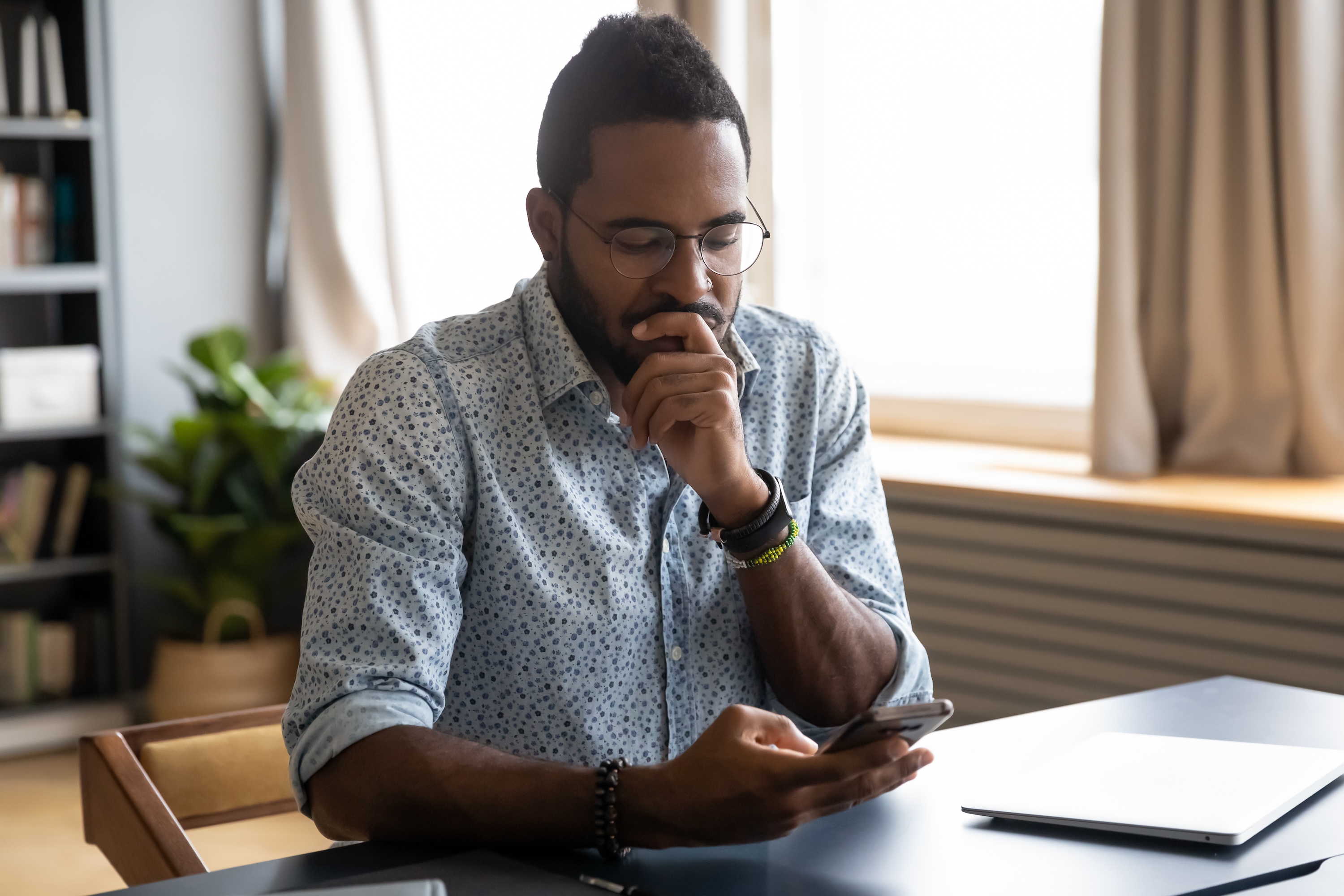 Confused African guy holding smart phone feels concerned thinking over received message.