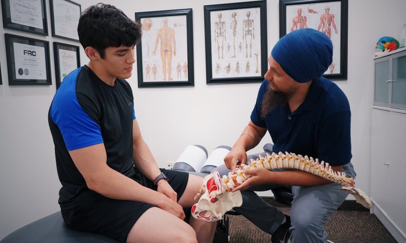 Doctor or healthcare professional discussing bone health and showing model of spine to patient sitting on bed in office