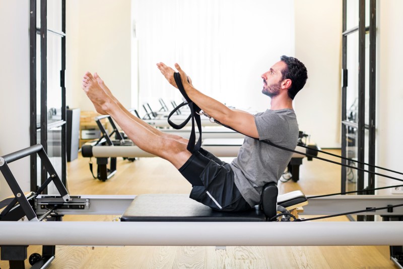 man doing pilates leg and arm stretch seated wooden floor