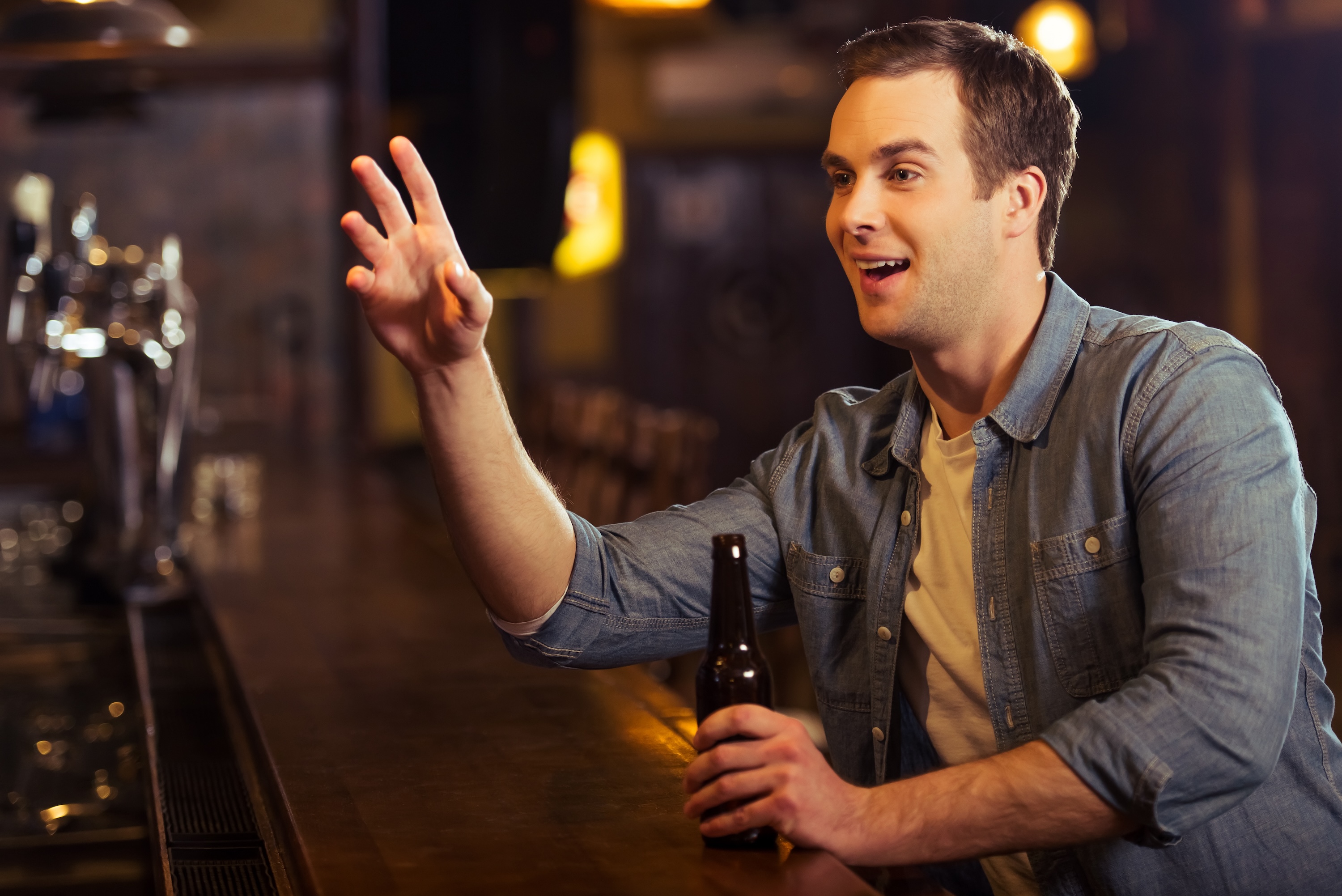 Young attractive man in casual clothes is smiling, holding a bottle of beer and making order while sitting at bar counter in pub