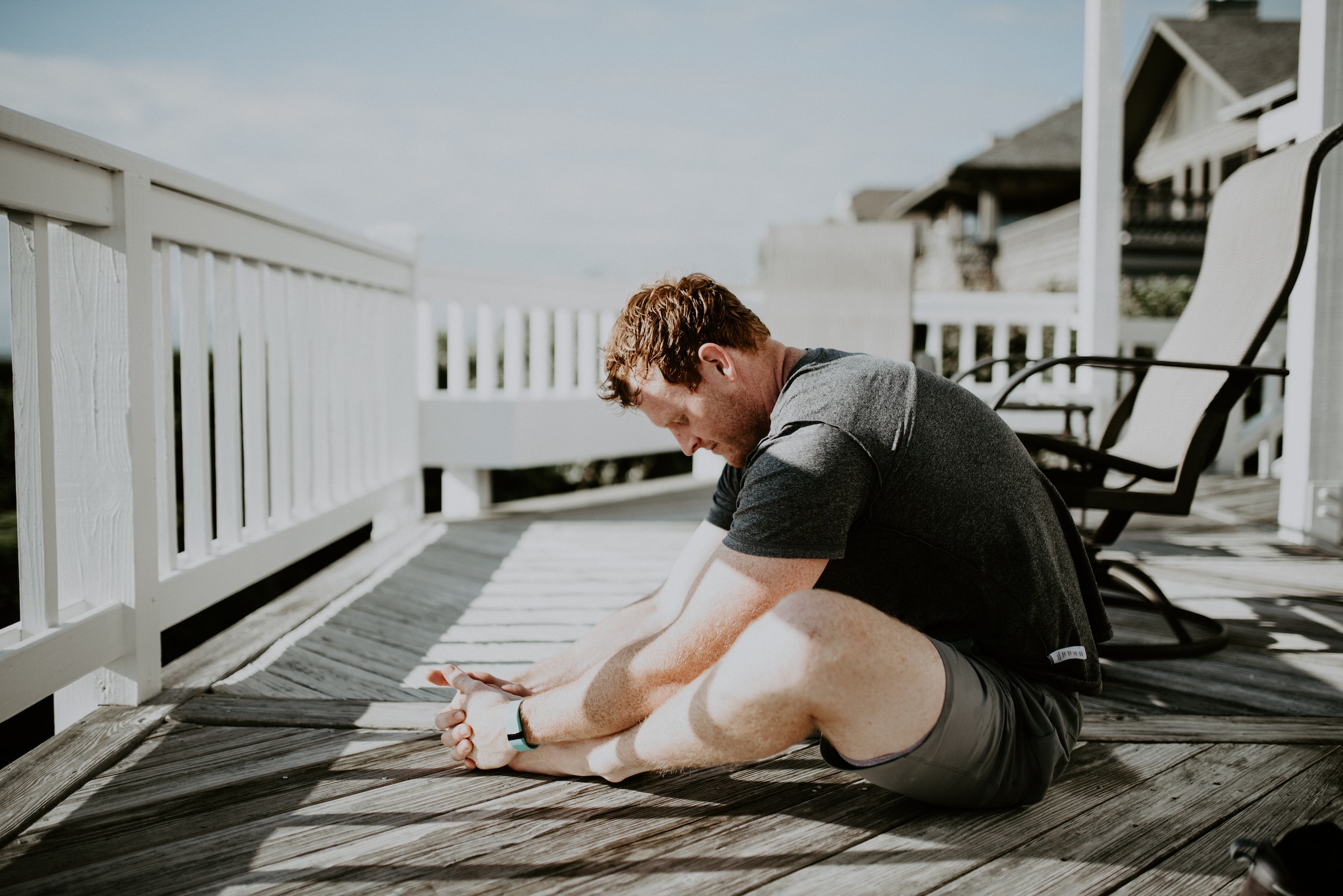 man sitting cross legged in a yoga pose on a porch outside by a white fence