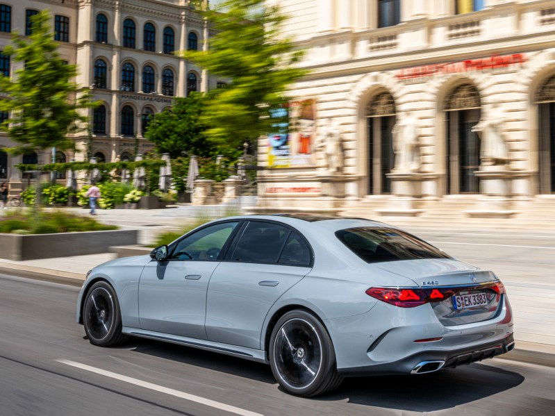 2024 Mercedes-Benz E-Class Sedan left rear three-quarter shot driving in city street with light stone buildings and a plaza with a few trees and people in the background.