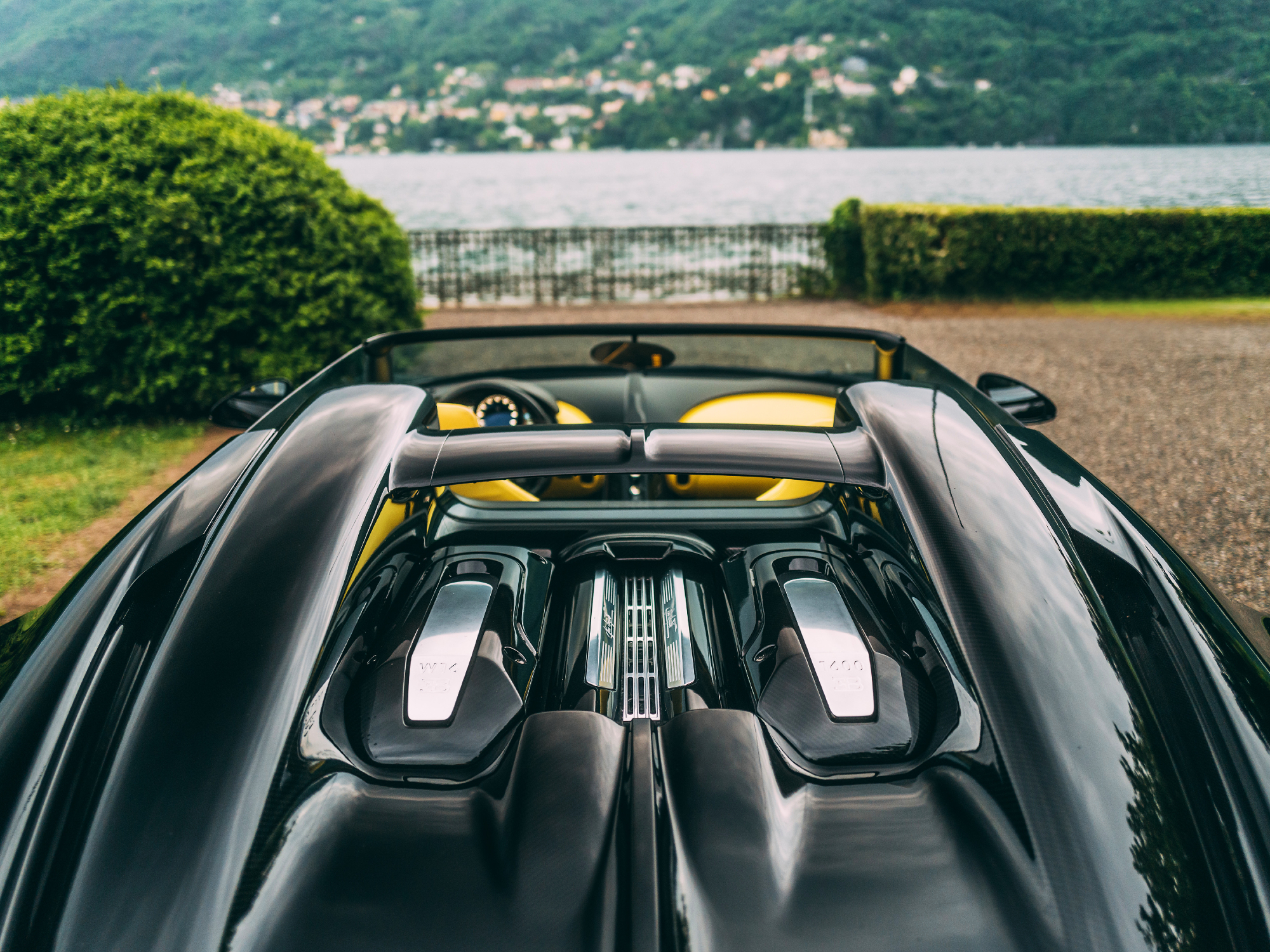 A view over the back of a black Bugatti W16 Mistral Roadster parked looking at a lake or harbor with a mountain side village on the other shore.