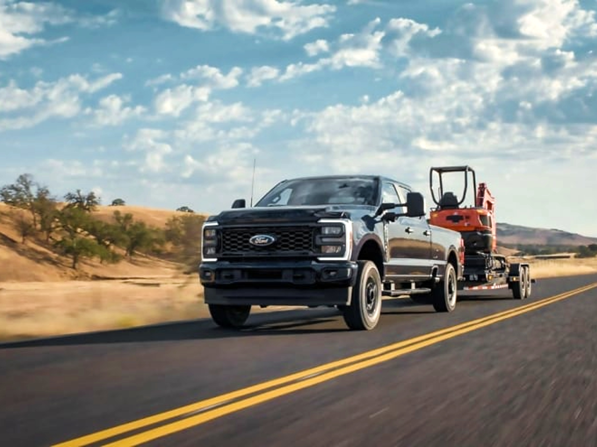 Ford F Series SuperDuty pickup truck towing a trailer carrying a piece of construction equipment on a highway.