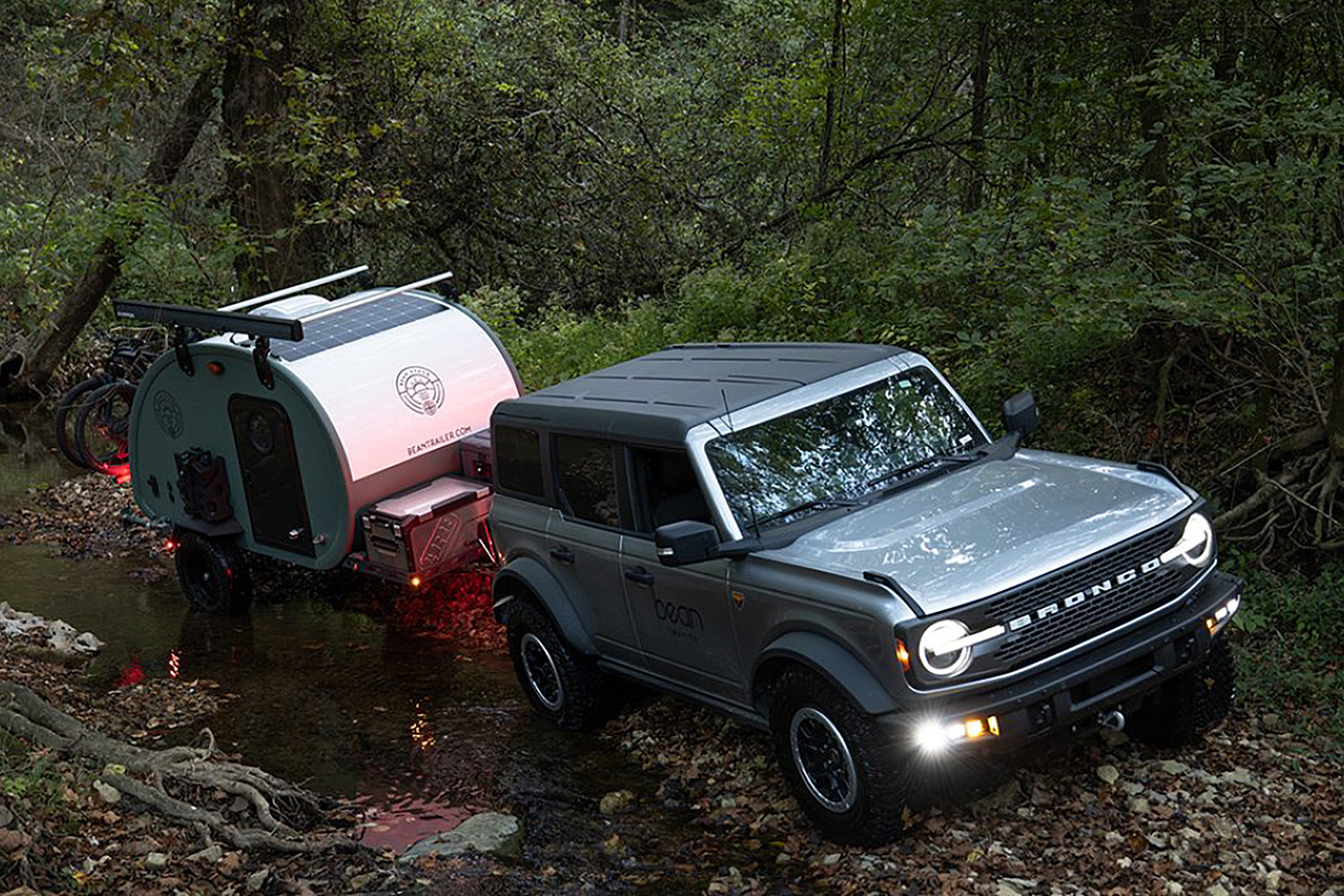 A Bean Stock 2.0 ultralight travel trailer being towed down a wooded trail by a new Ford Bronco.