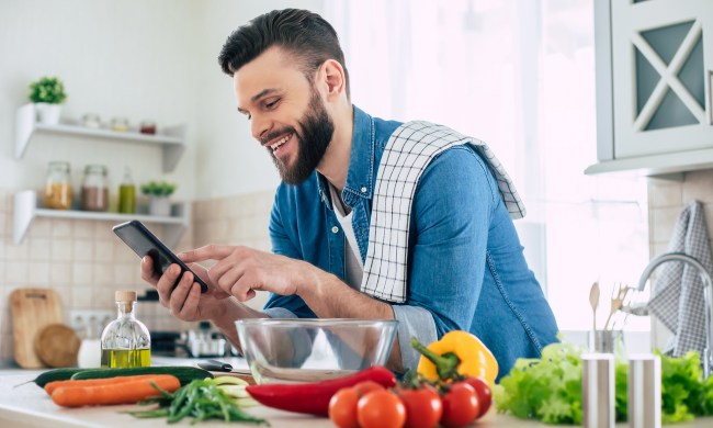 Happy bearded man is using his smartphone while he preparing a meal
