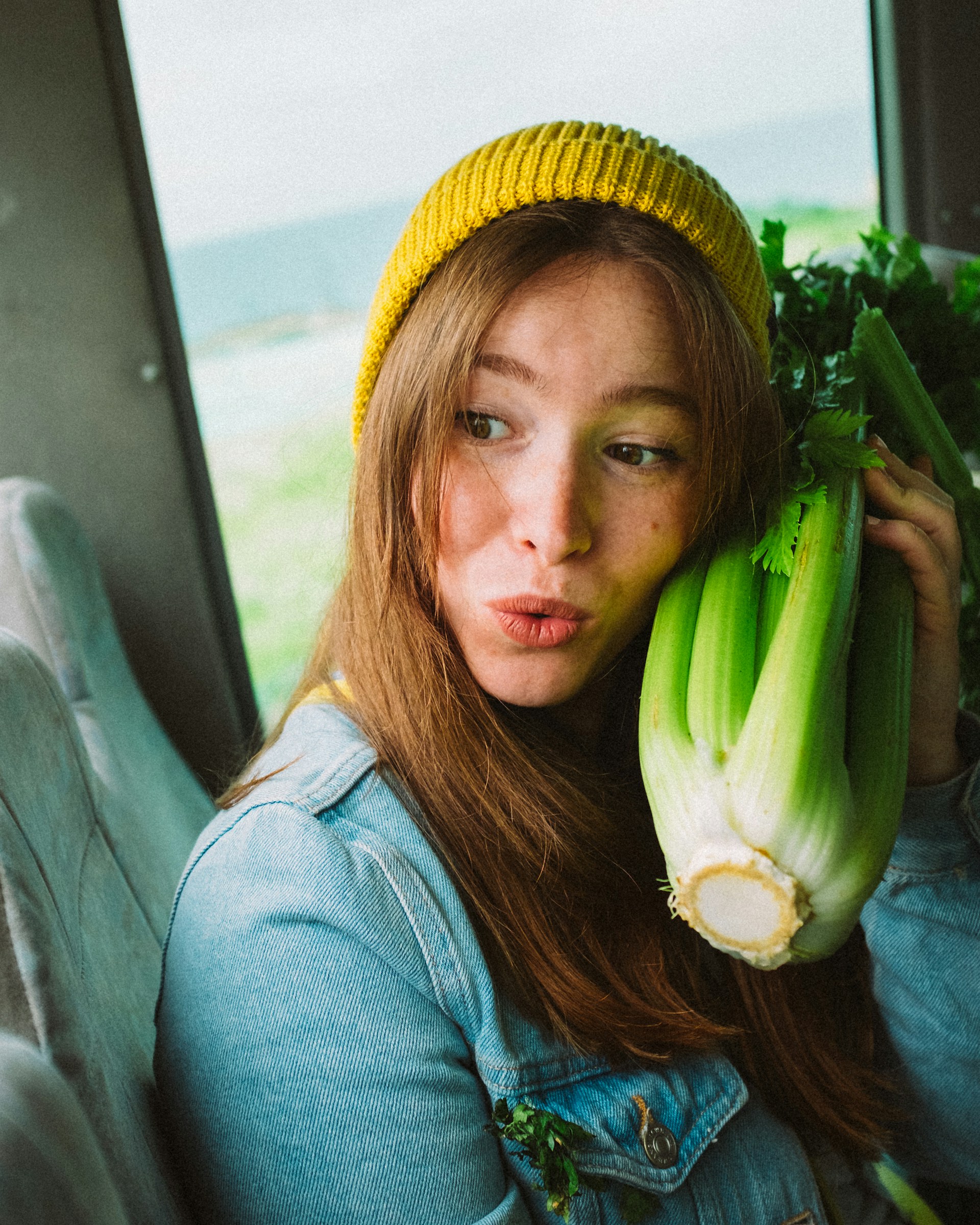 young woman wearing hat in car holding one bunch of green celery stalks close to her face