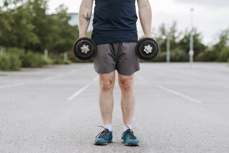 Person standing on asphalt holding a dumbbell in each hand.