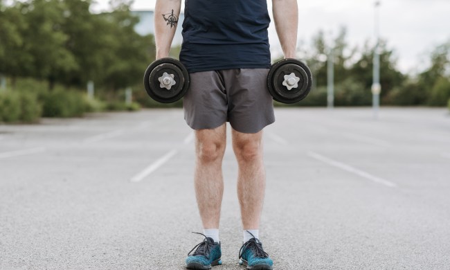 Person standing on asphalt holding a dumbbell in each hand.
