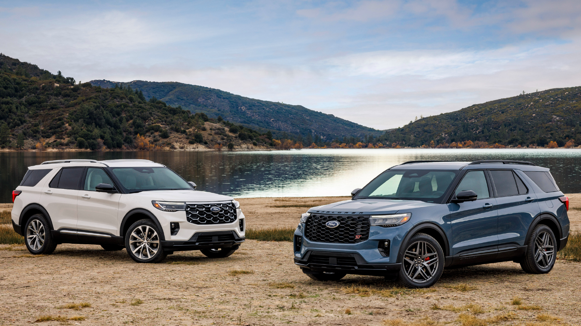 2025 Ford Explorer Platinum and 2025 Ford Explorer ST barked on stones by lakeside with moutains in the background.